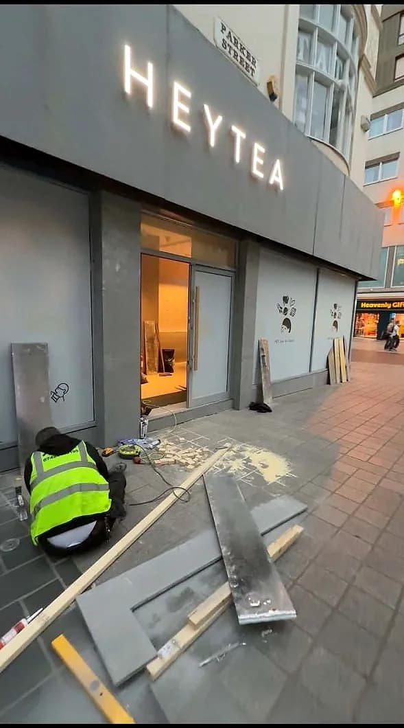 A MO Team fitter in hi-vis kneeling at the threshold of the HEYTEA unit at night, working on the new shopfront behind the hoarding