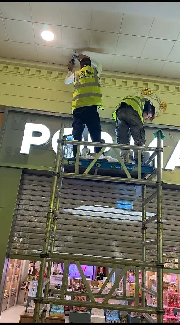 Two crew on a tower scaffold fitting the illuminated letters of the Pop Mart sign above the shutter