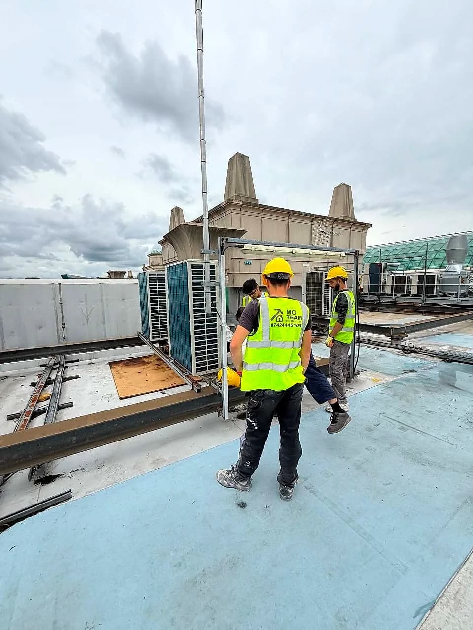 Two crew in hi-vis and hard hats installing air conditioning plant on a shopping centre roof under a grey sky