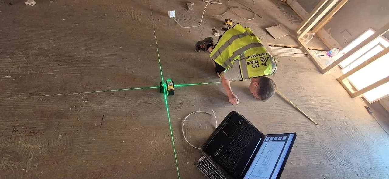 A crew member kneeling on a bare concrete floor with a green laser level and a set of drawings